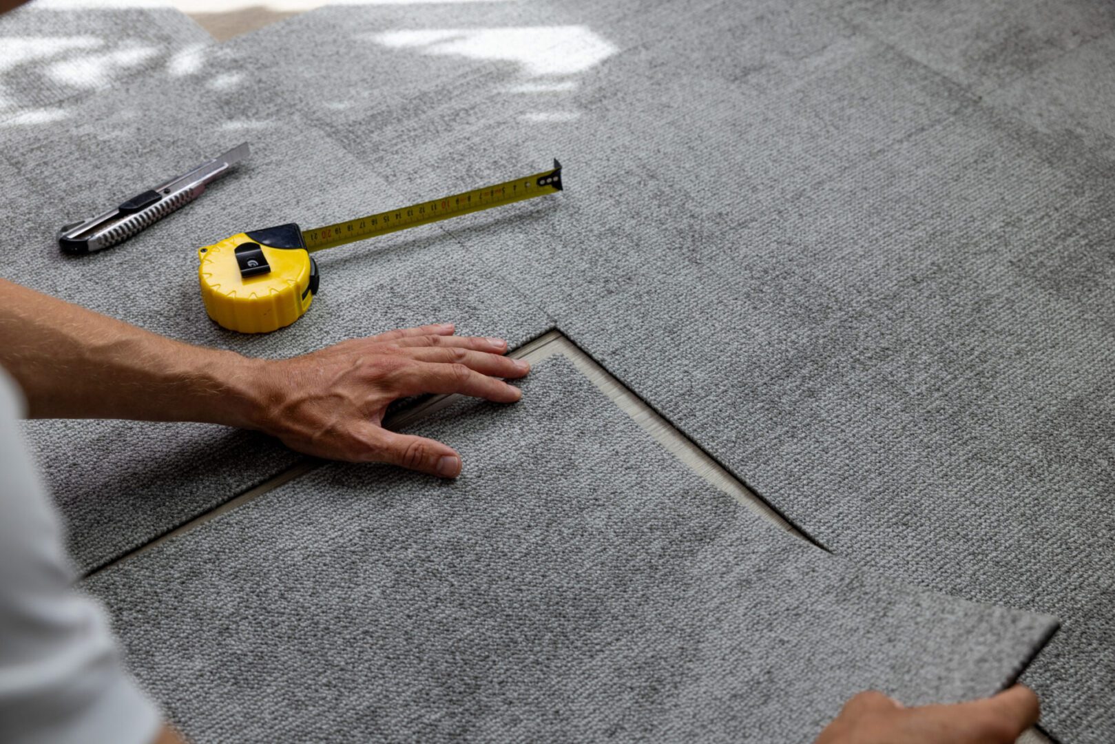 Hand installing gray carpet tiles, measuring tape.